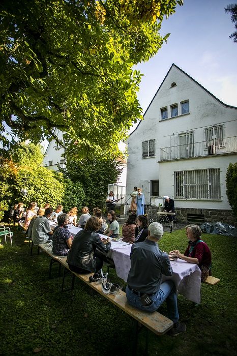 Eine Gruppe Theaterbesucher*innen sitzt im Garten eines Hauses an einr langen Tafel